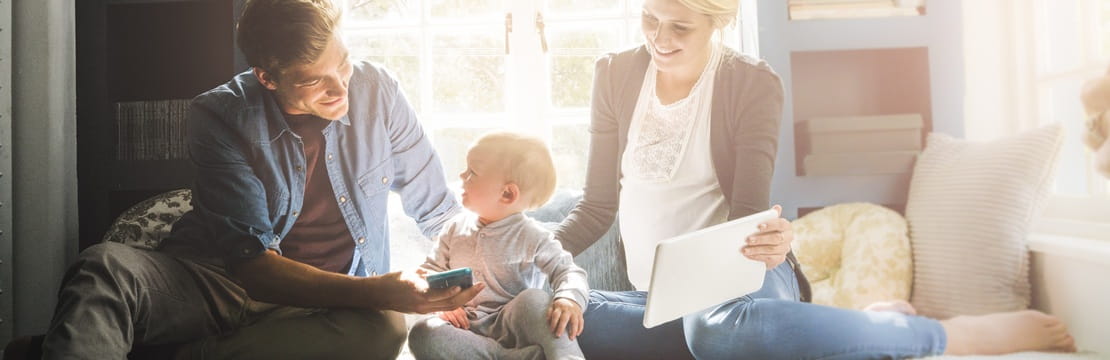 A young family sits together by a bright window, using a tablet and smartphone while discussing tax planning.